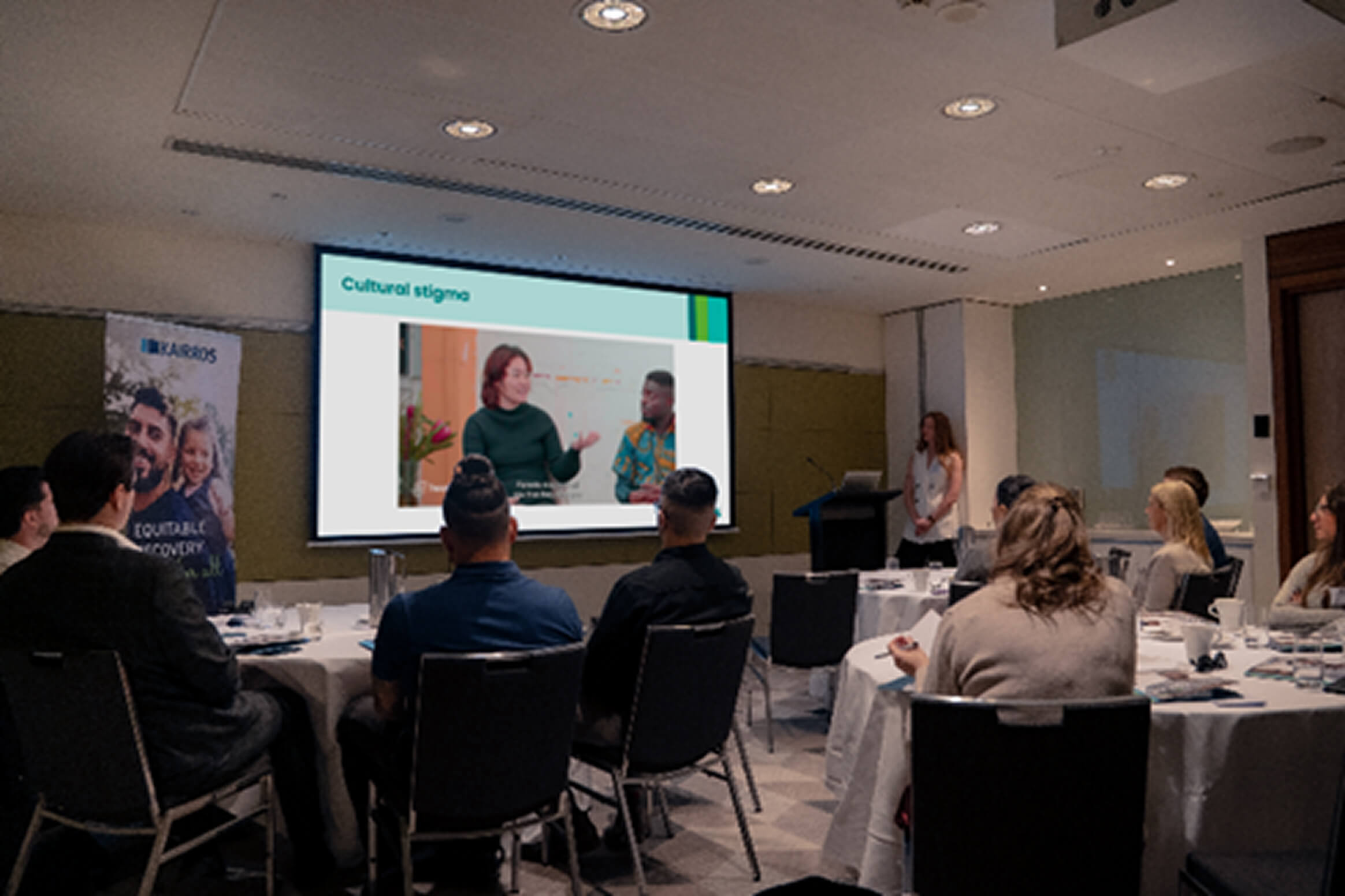 A projector in a room with conference tables and people sat at them
